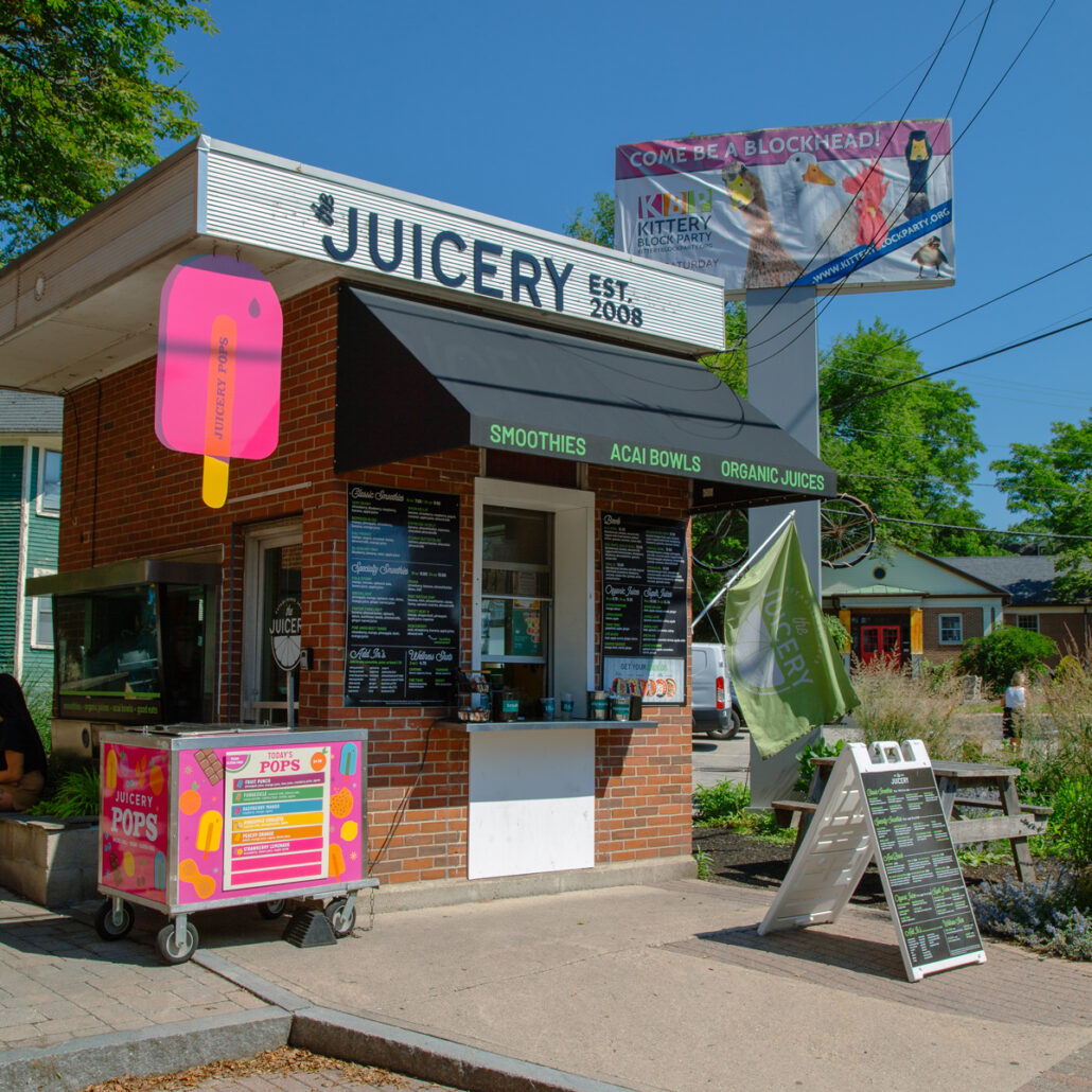 Exterior of The Juicery’s Kittery location with walk-up ordering window, menu boards, and colorful signage.