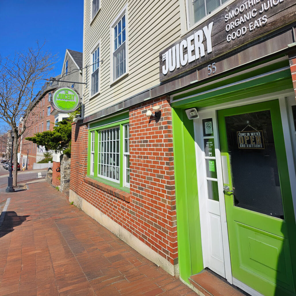 Exterior of The Juicery’s Portsmouth location with green trim, brick facade, and hanging logo sign.