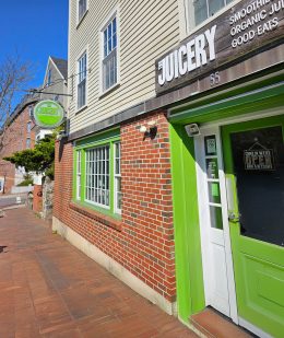 Exterior of The Juicery’s Portsmouth location with green trim, brick facade, and hanging logo sign.