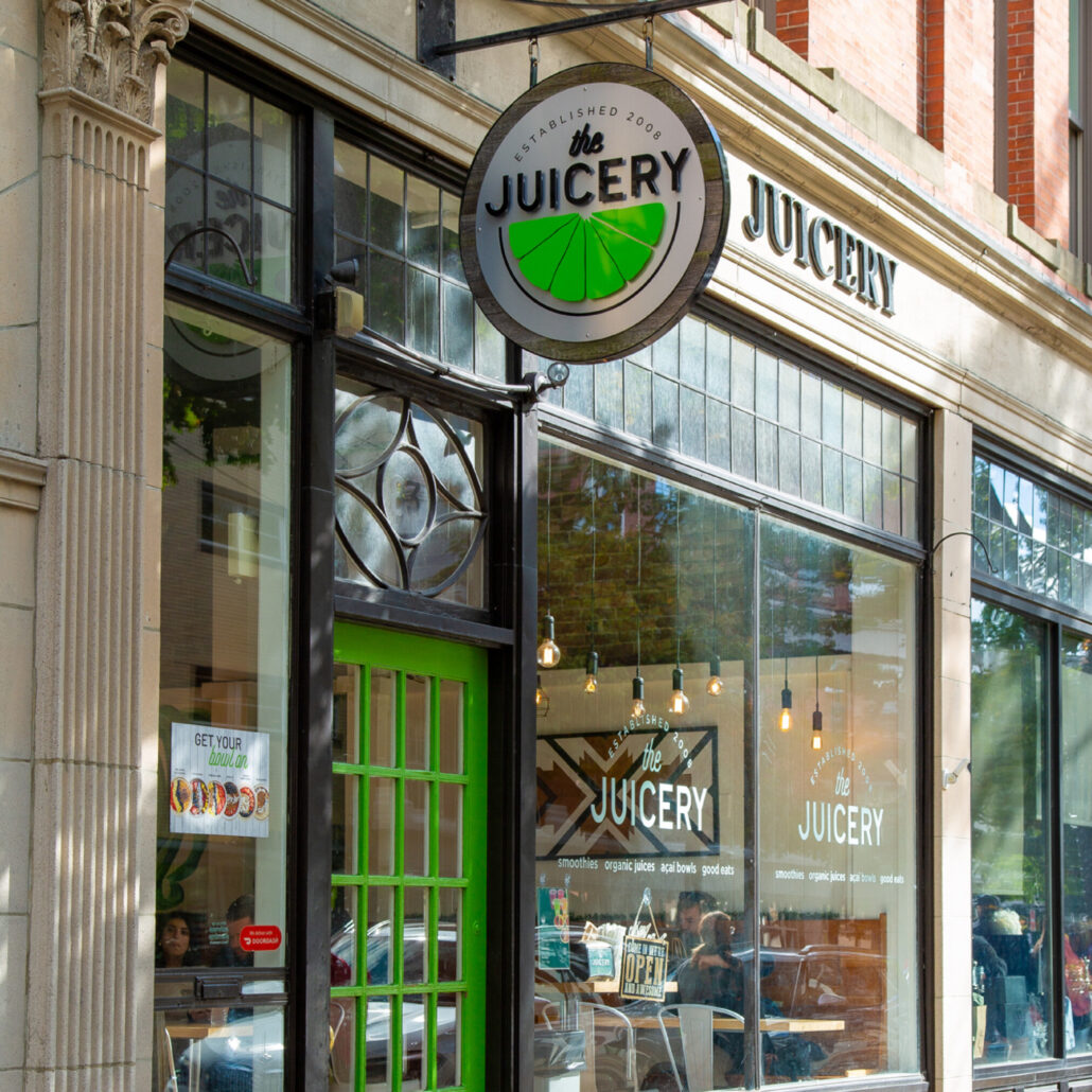 Exterior of The Juicery’s Salem location with green door, hanging logo sign, and large storefront windows.