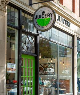 Exterior of The Juicery’s Salem location with green door, hanging logo sign, and large storefront windows.