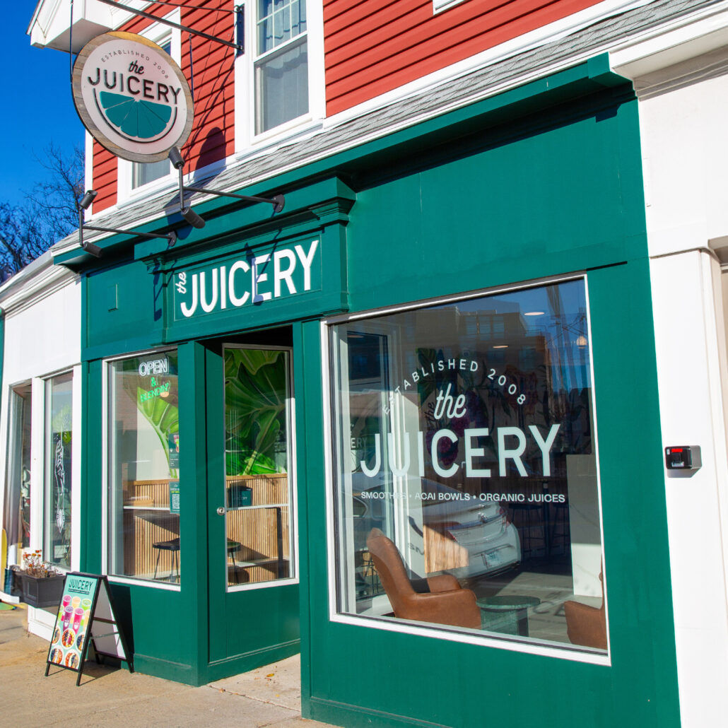 Exterior of The Juicery’s Dover location with green storefront, hanging logo sign, and large front windows.