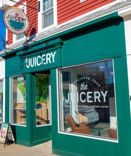 Exterior of The Juicery’s Dover location with green storefront, hanging logo sign, and large front windows.