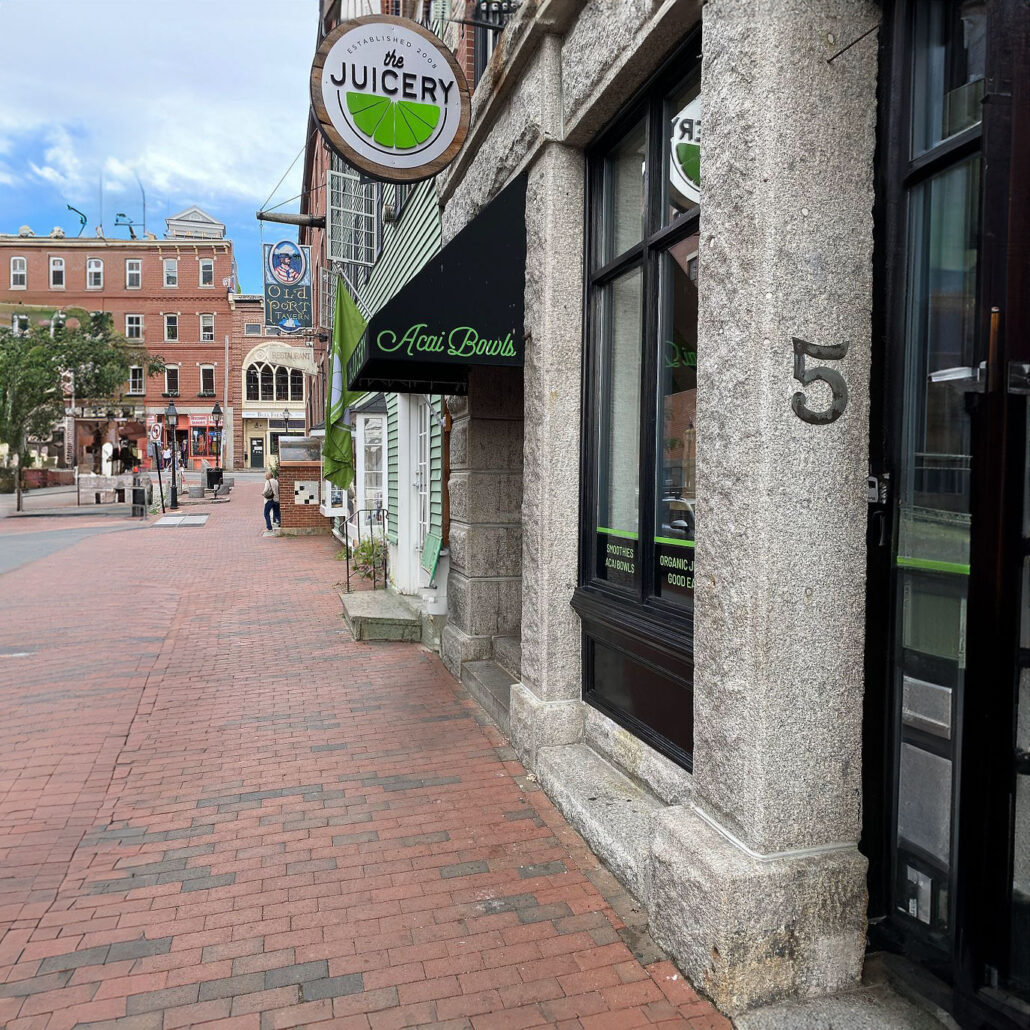 Exterior of The Juicery’s Portland location with green awning, hanging logo sign, and street-level entrance.