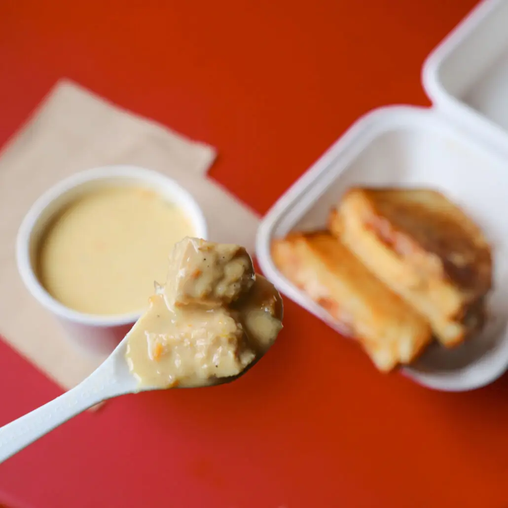 Spoonful of creamy soup held above a takeout container with grilled cheese in the background.